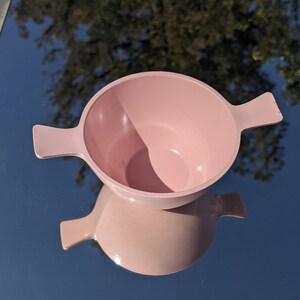 May include: A solid light pink bowl featuring two symmetrical, flat, horizontal handles extending from opposite sides. The bowl has a smooth, matte finish and a rounded interior basin, presented from a top-down perspective.