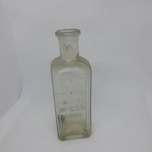 May include: A clear glass rectangular bottle with a narrow neck and a flared rim. The front of the bottle features embossed text that reads AN IDEAL TONIC, with additional faint lettering visible above and below the main phrase on the glass surface.