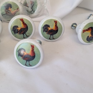 May include: A collection of round white ceramic cabinet knobs, each featuring a printed image of a rooster with black, orange, and red feathers against a light green background. The knobs include metal mounting screws attached to the back.