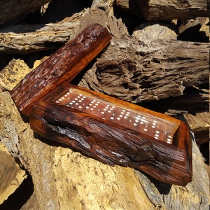 Puede incluir: Un juego de dominó de madera con un acabado de madera natural. El juego está alojado en una caja de madera rústica con una tapa con bisagras. La caja está hecha de una pieza gruesa de madera con una textura de corteza natural.