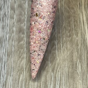 May include: A single, long, pink nail with glitter and small flecks of color. The nail is attached to a clear plastic stick. The nail is a light pink color with a glittery finish, and small flecks of gold, black, and purple.