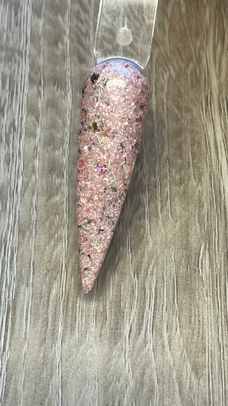 May include: A single, long, pink nail with glitter and small flecks of color. The nail is attached to a clear plastic stick. The nail is a light pink color with a glittery finish, and small flecks of gold, black, and purple.