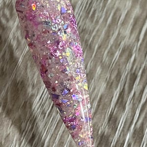 Puede incluir: Una muestra de u&ntilde;a transparente con un dise&ntilde;o de purpurina. La purpurina es una mezcla de copos rosas, morados e iridiscentes. La u&ntilde;a tiene forma de &oacute;valo largo y puntiagudo. La muestra se sujeta con un palito de pl&aacute;stico transparente.