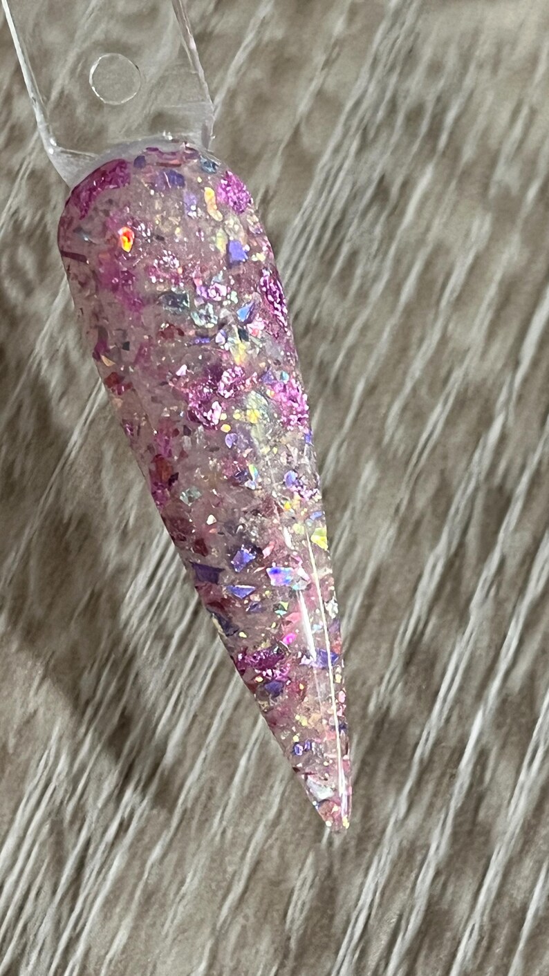 Puede incluir: Una muestra de u&ntilde;a transparente con un dise&ntilde;o de purpurina. La purpurina es una mezcla de copos rosas, morados e iridiscentes. La u&ntilde;a tiene forma de &oacute;valo largo y puntiagudo. La muestra se sujeta con un palito de pl&aacute;stico transparente.