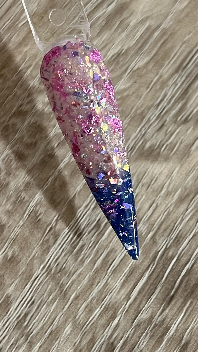 Puede incluir: Una muestra de u&ntilde;a larga y puntiaguda con un dise&ntilde;o degradado. La mitad superior es una mezcla de purpurina rosa y escamas iridiscentes, que transiciona a un azul profundo en la punta. La u&ntilde;a est&aacute; adherida a un palito de pl&aacute;stico transparente.