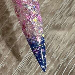 Puede incluir: Una u&ntilde;a larga y puntiaguda con un dise&ntilde;o degradado. La parte superior est&aacute; llena de purpurina rosa y morada, que pasa a un centro transparente y termina con una punta azul oscuro. La u&ntilde;a est&aacute; adherida a un palito de pl&aacute;stico transparente.