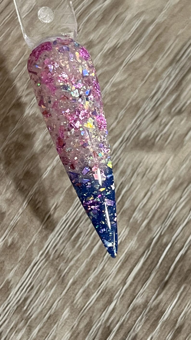 Puede incluir: Una u&ntilde;a larga y puntiaguda con un dise&ntilde;o degradado. La parte superior est&aacute; llena de purpurina rosa y morada, que pasa a un centro transparente y termina con una punta azul oscuro. La u&ntilde;a est&aacute; adherida a un palito de pl&aacute;stico transparente.
