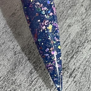 Puede incluir: Una muestra de u&ntilde;a con una base azul oscuro y escamas de purpurina iridiscente. La purpurina refleja varios colores, incluyendo morado, rosa y dorado. La u&ntilde;a es larga y puntiaguda, con un mango de pl&aacute;stico transparente.