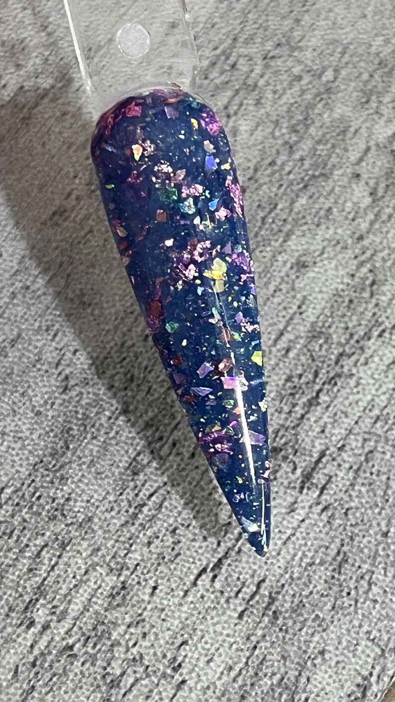 Puede incluir: Una muestra de u&ntilde;a con una base azul oscuro y escamas de purpurina iridiscente. La purpurina refleja varios colores, incluyendo morado, rosa y dorado. La u&ntilde;a es larga y puntiaguda, con un mango de pl&aacute;stico transparente.