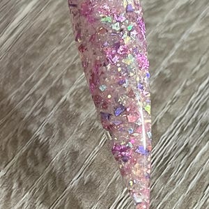 Puede incluir: Una muestra de u&ntilde;a transparente con un dise&ntilde;o de purpurina rosa. La purpurina incluye copos rosas, morados e iridiscentes de varios tama&ntilde;os y formas. La u&ntilde;a es larga y puntiaguda, con un mango de pl&aacute;stico transparente.