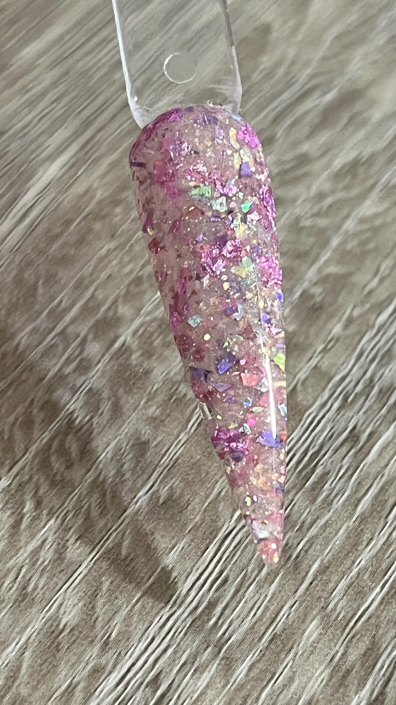 Puede incluir: Una muestra de u&ntilde;a transparente con un dise&ntilde;o de purpurina rosa. La purpurina incluye copos rosas, morados e iridiscentes de varios tama&ntilde;os y formas. La u&ntilde;a es larga y puntiaguda, con un mango de pl&aacute;stico transparente.