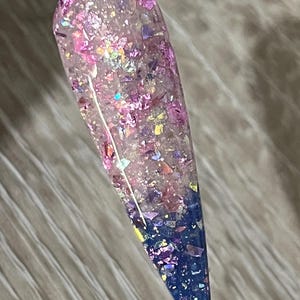 Puede incluir: Una u&ntilde;a larga y transparente con un dise&ntilde;o degradado. La mitad superior est&aacute; llena de purpurina rosa y morada iridiscente, que pasa a un azul profundo en la punta. La u&ntilde;a est&aacute; adherida a un palito de pl&aacute;stico transparente.