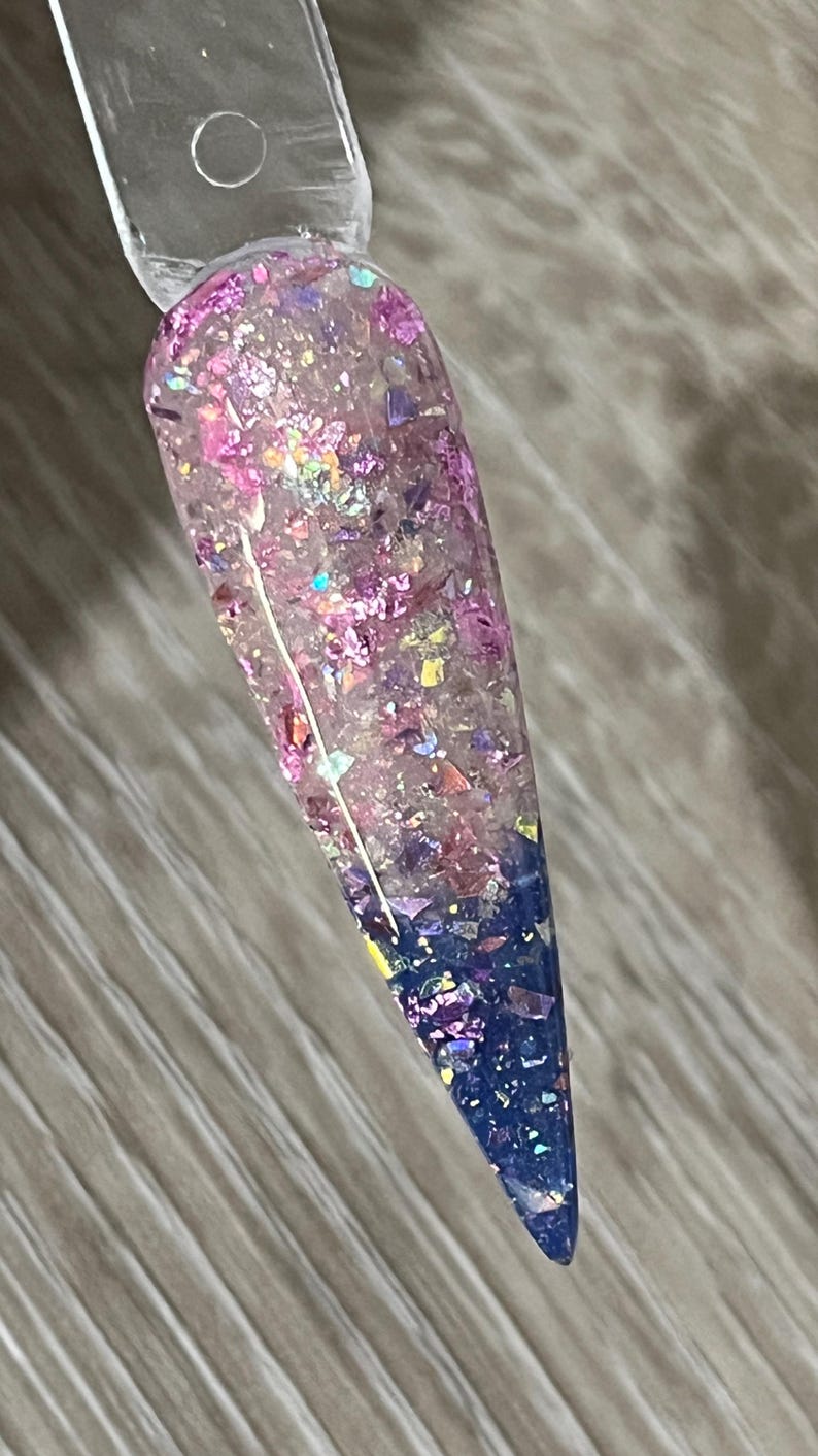 Puede incluir: Una u&ntilde;a larga y transparente con un dise&ntilde;o degradado. La mitad superior est&aacute; llena de purpurina rosa y morada iridiscente, que pasa a un azul profundo en la punta. La u&ntilde;a est&aacute; adherida a un palito de pl&aacute;stico transparente.