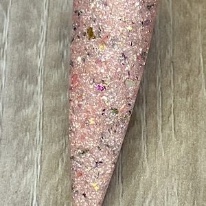 May include: A pink nail swatch with glitter and the text "Angel Kisses". The nail polish is a light pink color with iridescent glitter and small flecks of color. The nail is shaped into a long point.
