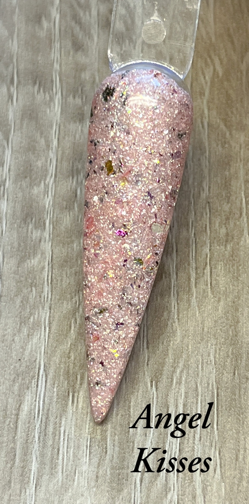 May include: A pink nail swatch with glitter and the text "Angel Kisses". The nail polish is a light pink color with iridescent glitter and small flecks of color. The nail is shaped into a long point.