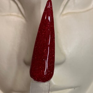 K&ouml;nnte beinhalten: Ein roter Glitzer-Nagellack mit einem klaren Applikator. Der Lack befindet sich in einem Fl&auml;schchen mit einem schwarzen Etikett, auf dem "B" steht.