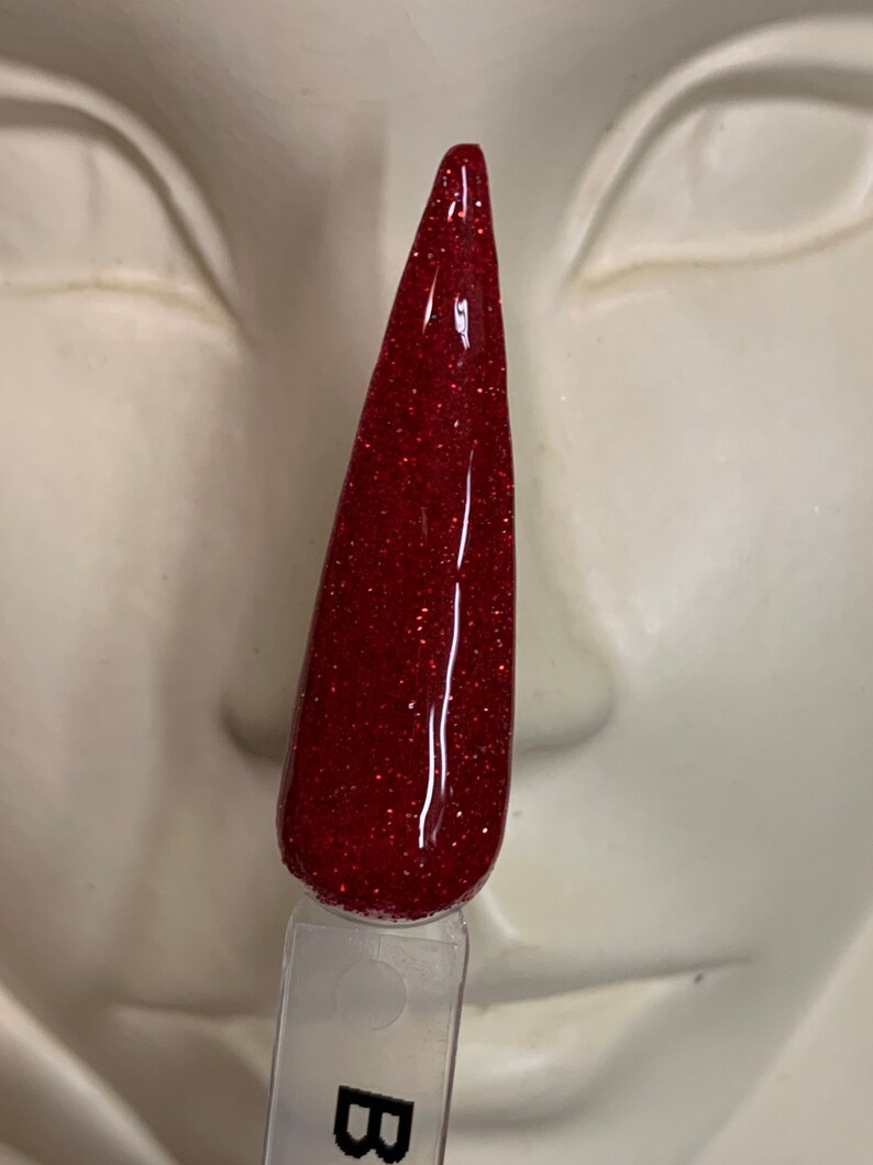K&ouml;nnte beinhalten: Ein roter Glitzer-Nagellack mit einem klaren Applikator. Der Lack befindet sich in einem Fl&auml;schchen mit einem schwarzen Etikett, auf dem "B" steht.