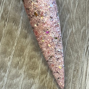May include: A single, long, pointed nail with a light pink base color. The nail is covered in a glittery mix of silver, gold, and iridescent particles. The nail is attached to a clear plastic stick.