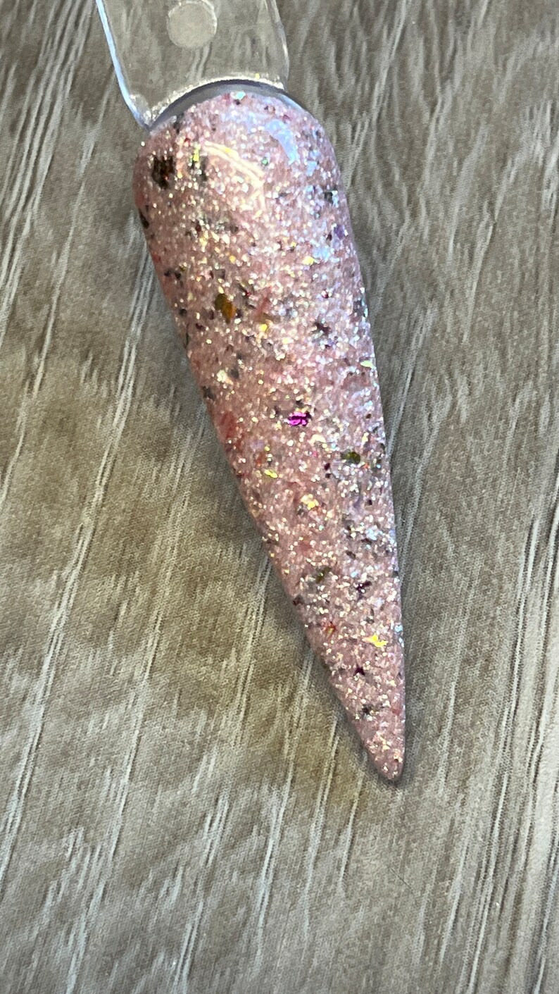 May include: A single, long, pointed nail with a light pink base color. The nail is covered in a glittery mix of silver, gold, and iridescent particles. The nail is attached to a clear plastic stick.