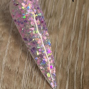 May include: A clear, pointed nail swatch stick featuring a dense mix of pink, purple, and iridescent hexagonal and irregular glitter flakes. The word "Dreamspell" is printed in black text next to the swatch, which displays a multi-dimensional, reflective finish with various sizes of shimmering particles embedded throughout the clear base.