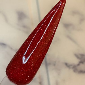 K&ouml;nnte beinhalten: Nahaufnahme eines roten Nagellacks mit schimmerndem Finish. Der Lack ist auf einen einzelnen Nagel aufgetragen, der in eine lange, spitze ovale Form gebracht wurde.