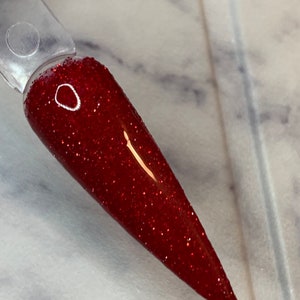 K&ouml;nnte beinhalten: Nahaufnahme eines roten Glitzer-Nagellacks auf einem klaren Applikatorstab. Der Lack hat eine feine Glitzer-Oberfl&auml;che und ist in einem tiefen Rotton.