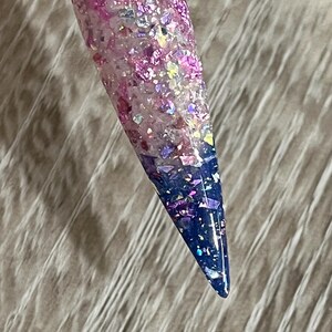 Puede incluir: Una u&ntilde;a larga y puntiaguda con un dise&ntilde;o degradado. La u&ntilde;a hace la transici&oacute;n de una base rosa claro a una punta azul oscuro, con purpurina y copos iridiscentes. La u&ntilde;a est&aacute; adherida a un palito de pl&aacute;stico transparente.