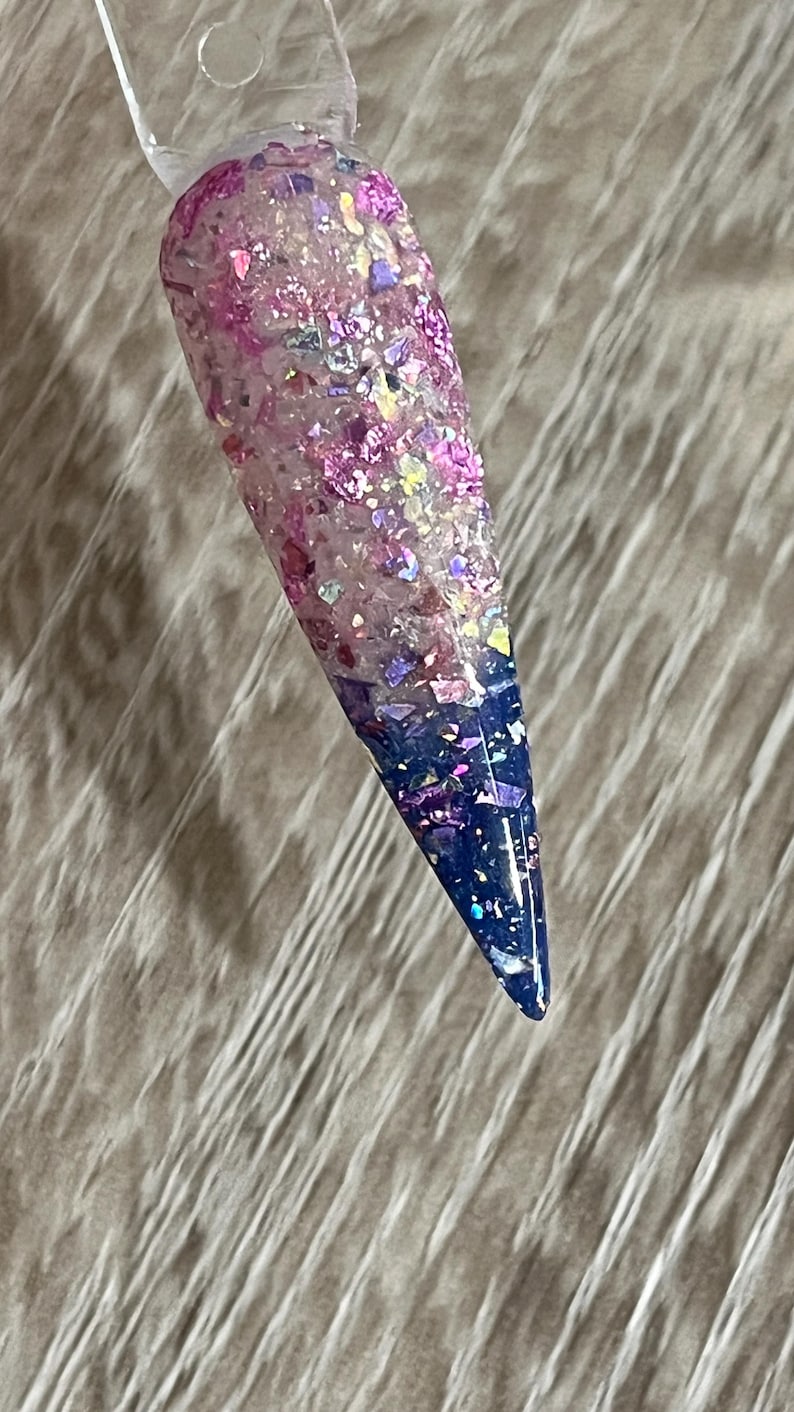 Puede incluir: Una u&ntilde;a larga y puntiaguda con un dise&ntilde;o degradado. La u&ntilde;a hace la transici&oacute;n de una base rosa claro a una punta azul oscuro, con purpurina y copos iridiscentes. La u&ntilde;a est&aacute; adherida a un palito de pl&aacute;stico transparente.