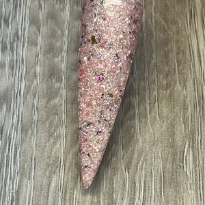 May include: A nail swatch featuring a pink glitter nail polish with flecks of black, gold, and iridescent glitter. The nail polish is applied to a clear plastic stick, showcasing the color and texture of the product.