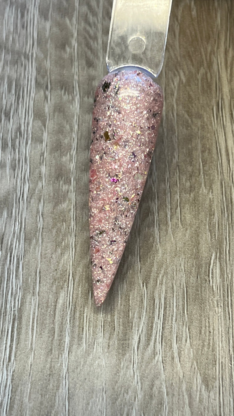 May include: A nail swatch featuring a pink glitter nail polish with flecks of black, gold, and iridescent glitter. The nail polish is applied to a clear plastic stick, showcasing the color and texture of the product.