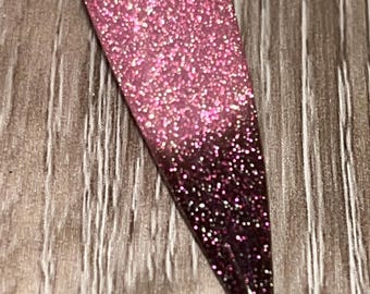 May include: A pink and burgundy glitter nail polish with a gradient effect. The polish is applied to a single nail.