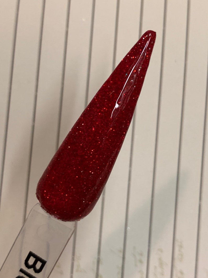 K&ouml;nnte beinhalten: Nahaufnahme eines roten Glitzer-Nagellacks auf einem durchsichtigen Applikatorstab. Der Lack hat eine feine Glitzer-Oberfl&auml;che und ist tiefrot.