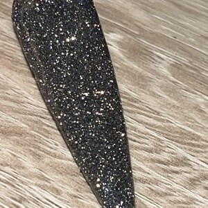May include: A close-up of a dark gray nail polish with a sparkly finish. The polish is applied to a long, pointed nail.