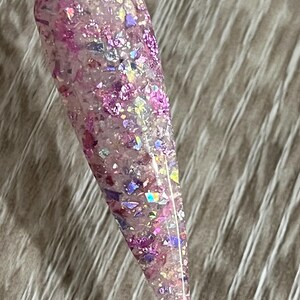 Puede incluir: Una u&ntilde;a larga y transparente con un dise&ntilde;o de purpurina rosa y morada. La purpurina incluye copos iridiscentes y trozos m&aacute;s grandes, creando un efecto brillante. La u&ntilde;a est&aacute; sujeta por un palito de pl&aacute;stico transparente.