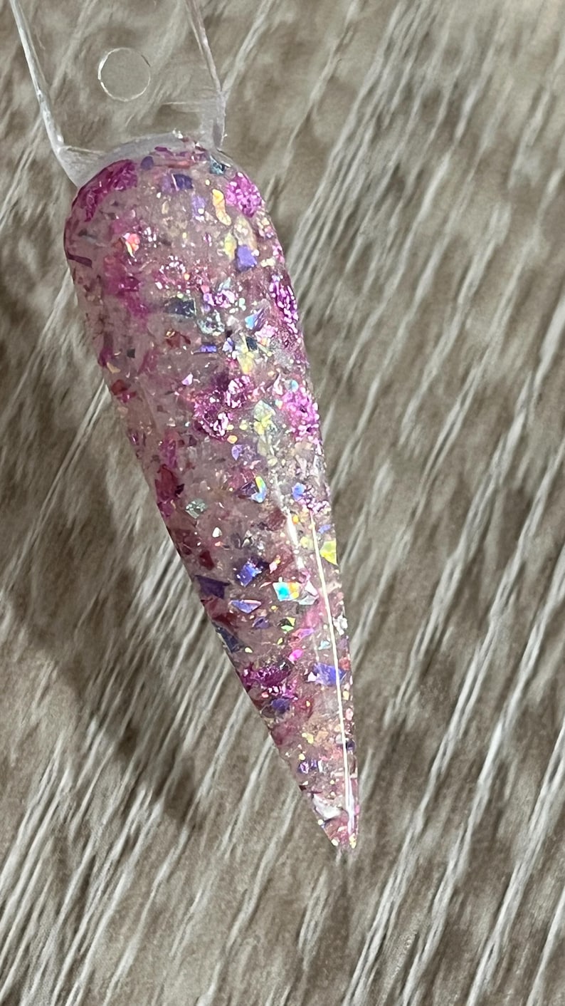 Puede incluir: Una u&ntilde;a larga y transparente con un dise&ntilde;o de purpurina rosa y morada. La purpurina incluye copos iridiscentes y trozos m&aacute;s grandes, creando un efecto brillante. La u&ntilde;a est&aacute; sujeta por un palito de pl&aacute;stico transparente.