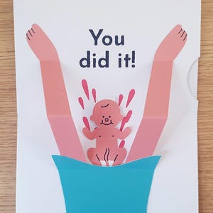 Könnte beinhalten: Eine weiße Grußkarte mit einem Pop-up-Design aus zwei rosa Beinen, die von einer türkisfarbenen Basis nach oben ragen. Zwischen den Beinen befindet sich eine kleine rosa Baby-Illustration mit roten Spritzern und dem dunkelblauen Text You did it!, der über der Babyfigur gedruckt ist.