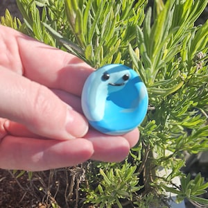 Könnte beinhalten: Eine kleine, blaue und weiße Keramikfigur mit einem lächelnden Gesicht. Die Figur ist rund und dicklich geformt und hat einen weißen Umriss.