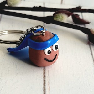 May include: A brown clay keyring with a blue headband and googly eyes. The keyring is shaped like a person with a smiling face.