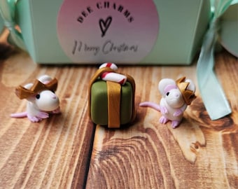 May include: A festive scene featuring a mint green gift box with a pink label that reads "Dee Charms" and "Merry Christmas." Three miniature mice wearing crowns and a wrapped gift are arranged on a wooden surface.