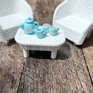 May include: A miniature white wicker-style furniture set consisting of two chairs and a rectangular table. On the table sits a light blue teapot, a matching creamer, and two small cups, all decorated with white and yellow daisy flower patterns.