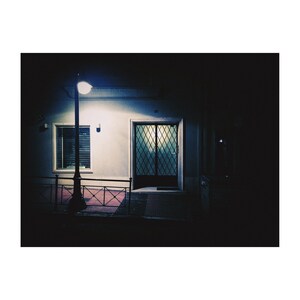 May include: A dark, nighttime view of a building with a single light illuminating the front door. The door is made of glass with a metal grate and is closed. There is a window to the left of the door and a street lamp in front of the building.