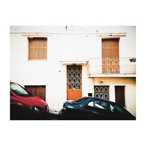 May include: A red car parked in front of a white building with a brown door and windows. A green car is parked behind the red car.
