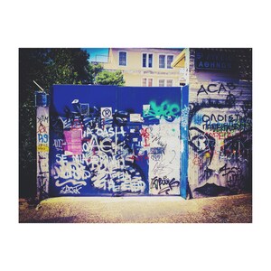 May include: A blue metal gate with graffiti art. The gate is covered in colorful spray paint with words and symbols. The gate is in front of a building with a white wall and a sign that says "5th Avenue Athens".