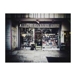 May include: A storefront with a sign that reads "ΓΡΑΦΕΙΟΝ ΚΑΛΟΥ ΤΥΠΟΥ" in gold letters. The storefront has large windows displaying rows of books and other items.