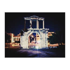 May include: A white stone archway with a large opening in the middle. The archway is lit up at night and there are buildings and streetlights in the background.