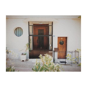 May include: A white building with a brown wooden door with a black metal gate. The door has a small round window with a black frame. There are plants in front of the door and a white planter with a plant in it.