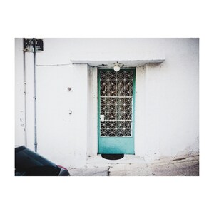 May include: A teal blue door with a white metal grate pattern and a light fixture above. The door is set in a white wall with a gray pipe running down the side.