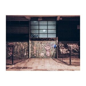 May include: A brick wall with graffiti art featuring a hand with an arrow pointing to the right, geometric shapes, and a face. The wall is painted in shades of pink, blue, green, and yellow. The graffiti is surrounded by a black metal fence.