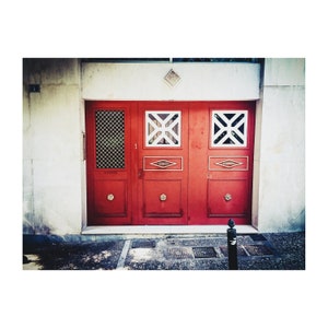 May include: A red double door with three windows, each with a white geometric design. The door is set in a white wall with a small diamond-shaped design above it.