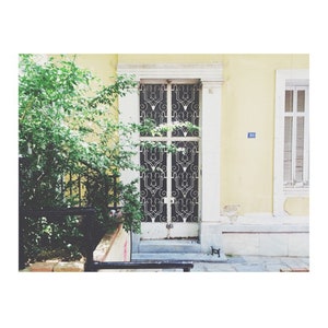 May include: A white door with a black metal grate design. The door is on a yellow building with a blue house number.